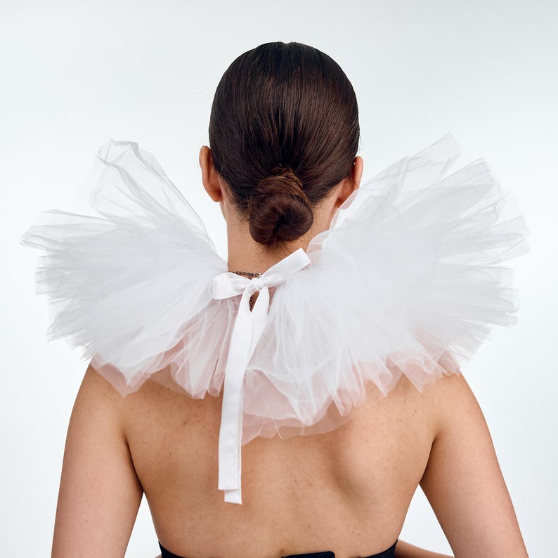 May include: A white tulle collar with a ruffled design, tied at the back with a white satin ribbon. The collar is worn around the neck, creating a dramatic and elegant look. The person's hair is styled in a bun.