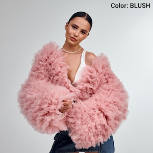 Könnte beinhalten: Eine blushfarbene Tülljacke mit Rüschendesign. Die Jacke hat lange Ärmel und eine voluminöse Silhouette. Der Text "Color: BLUSH" befindet sich in der oberen rechten Ecke. Das Model trägt ein weißes Top und eine Perlenkette.