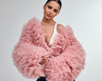 May include: A blush-colored tulle jacket with a ruffled design. The jacket has long sleeves and a voluminous silhouette. The text "Color: BLUSH" is in the upper right corner. The model is wearing a white top and a pearl necklace.