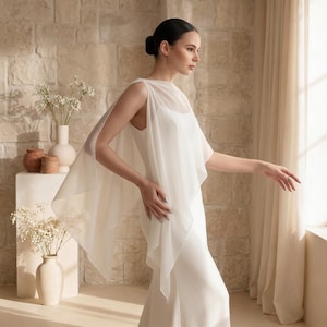 May include: A floor-length white gown featuring a sleeveless bodice and a sheer, flowing chiffon cape overlay that drapes over the shoulders and arms. The dress has a simple, clean silhouette with a round neckline and a smooth, solid fabric finish.