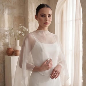 May include: A white sleeveless dress featuring a sheer, lightweight organza capelet overlay. The capelet has a high, rounded neckline and drapes loosely over the shoulders and arms, creating a soft, layered effect over the solid white bodice.