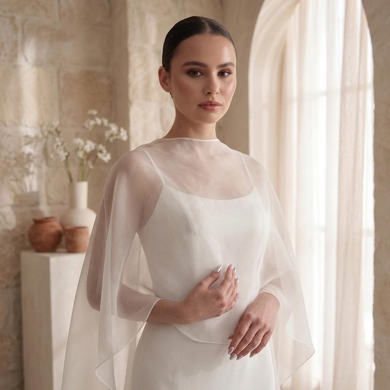 May include: A white sleeveless dress featuring a sheer, lightweight organza capelet overlay. The capelet has a high, rounded neckline and drapes loosely over the shoulders and arms, creating a soft, layered effect over the solid white bodice.