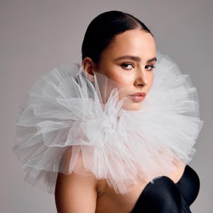 May include: A person is wearing a voluminous white tulle collar, the main feature of the image. The collar is layered and stands out against a black top. The person has dark hair and is looking towards the camera.