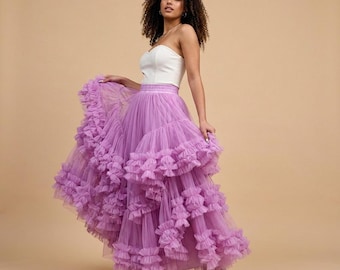 May include: A woman wearing a white strapless top and a layered, ruffled lavender tulle skirt. The skirt has multiple tiers of cascading ruffles, creating a voluminous silhouette. The outfit is paired with silver sandals.