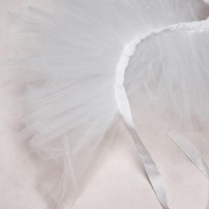 May include: A close-up of a white tutu skirt. The skirt is made of layers of sheer, lightweight fabric. The waistband is a solid white, and a white ribbon is attached. The tutu is likely for dance or costume.