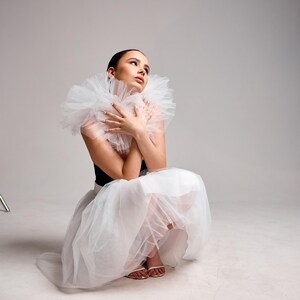 May include: A person wearing a black top and white tulle skirt, with a large white ruffled collar. The person is wearing white sandals and is sitting on a white surface. The background is a plain grey.