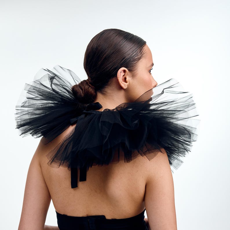 May include: A black tulle collar with multiple layers, creating a voluminous effect. The collar is tied at the back with black ribbons. The model is wearing a black top, and the background is white.