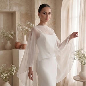May include: A white sleeveless floor-length dress featuring a sheer, lightweight chiffon capelet overlay that drapes over the shoulders and arms, creating a flowing silhouette with a high neckline and a clean, minimalist aesthetic.
