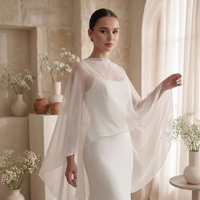 May include: A white sleeveless floor-length dress featuring a sheer, lightweight chiffon capelet overlay that drapes over the shoulders and arms, creating a flowing silhouette with a high neckline and a clean, minimalist aesthetic.