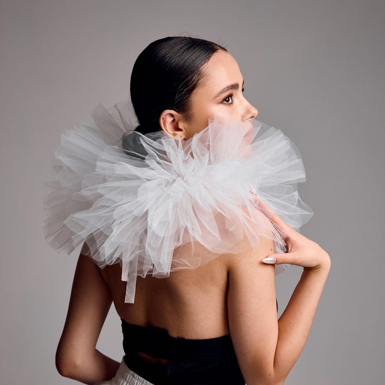 May include: A layered white tulle collar, offering a voluminous appearance. The collar is worn over a black strapless top. The image is set against a neutral grey backdrop, emphasizing the collar's texture and design.