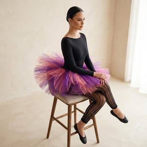 May include: A person wearing a black long-sleeve leotard and a layered tutu skirt in shades of purple and pink. The person is also wearing black leggings and black ballet flats. The person is sitting on a wooden stool.