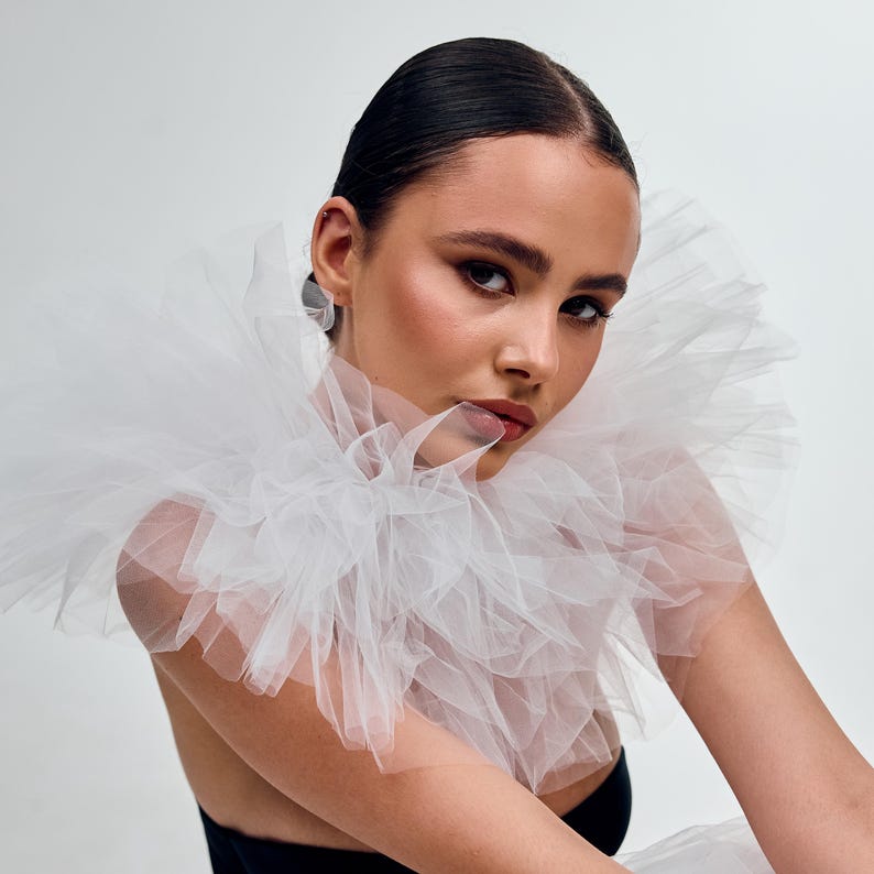 May include: A woman wearing a white tulle collar and a black top. The collar is layered and ruffled, creating a voluminous effect. The woman has dark hair pulled back and is wearing silver earrings. The background is a plain white.