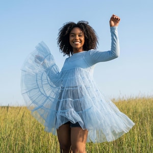 May include: A light blue long-sleeved dress with a tiered, flowing skirt made of tulle. The dress is worn outdoors in a field of tall grass under a clear blue sky. The model is smiling and has their arm raised.
