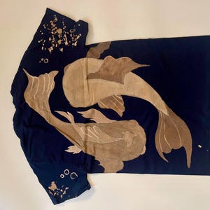 May include: Navy blue t-shirt featuring a koi fish design in shades of tan. The fish are swimming in opposite directions. The shirt has a unique, artistic design, with a splash of tan and white paint on the left shoulder.