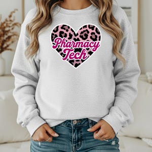 Puede incluir: Sudadera gris claro con un diseño de corazón de leopardo rosa y negro y la inscripción "Pharmacy Tech". La sudadera se combina con unos vaqueros azules.