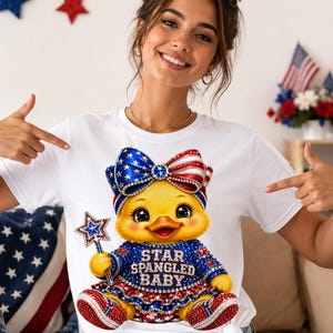 Puede incluir: Una camiseta blanca de manga corta con el gráfico de un patito amarillo que lleva un lazo rojo, blanco y azul y un conjunto a juego con el texto STAR SPANGLED BABY. El patito sostiene una varita en forma de estrella, todo decorado con pedrería.