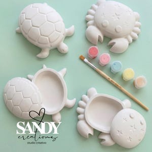 May include: White ceramic turtle and crab shaped paint-your-own figurines. The turtle and crab figurines are shown with their lids open and closed. The image also includes a paintbrush and small paint pots in various colors.