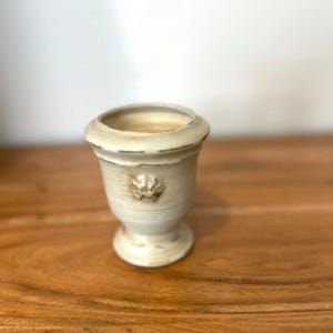 Puede incluir: Una pequeña urna decorativa de color crema con un acabado envejecido. Presenta un diseño floral detallado y una base de pedestal. El interior es de un color crema más claro. La urna está sobre una superficie de madera.