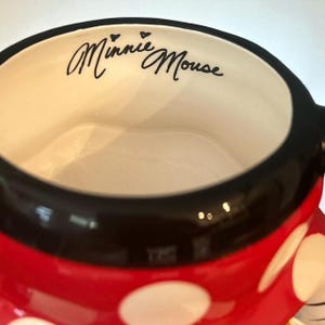 May include: A ceramic mug with a red base and white polka dots. The rim is black, and the interior is off-white. The words "Minnie Mouse" are written in black script inside the mug.