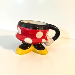 May include: A ceramic Minnie Mouse-themed coffee mug. The mug features a red body with white polka dots, black handle and rim, and yellow shoes. The mug is designed to look like Minnie Mouse's iconic outfit.