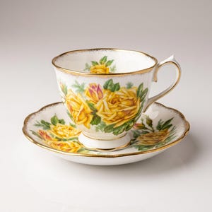 May include: White teacup and saucer set with a yellow rose floral design and gold trim. The teacup has a handle and sits on a matching saucer. The set is likely made of porcelain.