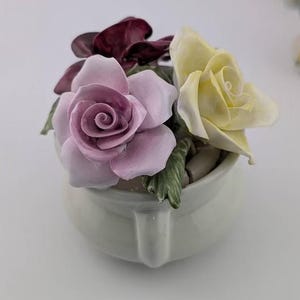 Puede incluir: Un arreglo floral de cerámica con una rosa rosa claro, una rosa amarilla y hojas de color rojo oscuro. Las flores están en una pequeña maceta blanca con asa. El arreglo está sobre una superficie blanca.