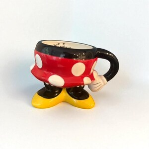 May include: A ceramic mug shaped like Minnie Mouse, featuring a red and white polka-dotted skirt, black handle and rim, and yellow shoes. The mug has a white glove-shaped hand holding the handle. The mug is designed to stand upright.