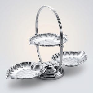 Puede incluir: Una bandeja de servir de tres niveles con asa. La bandeja es de metal plateado brillante y presenta tres platos en forma de hoja con una textura martillada. El asa se arquea sobre la parte superior, conectándose a la base.