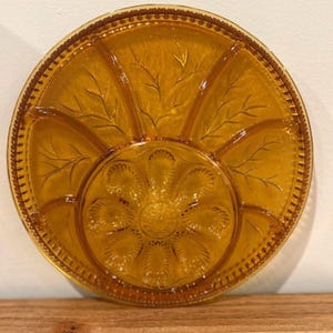 Peut inclure: Un plateau de service rond en verre ambré doté d'un compartiment central circulaire avec un motif floral, entouré de quatre sections extérieures décorées de motifs de feuilles en relief et d'un bord perlé sur le pourtour du plat.