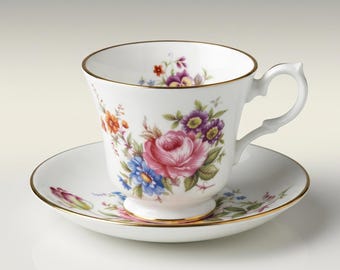 Puede incluir: Un juego de taza de té y platillo de porcelana blanca con un colorido estampado floral de rosas rosas, flores azules y flores moradas. El borde tanto de la taza como del platillo tiene un fino acabado en oro y la taza presenta un asa curva.