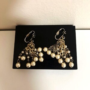 May include: A pair of dangling earrings with a mix of gold-toned metal and faux pearls. The earrings feature a cluster of small pearls hanging from a decorative metal design. The earrings are displayed on a black surface.