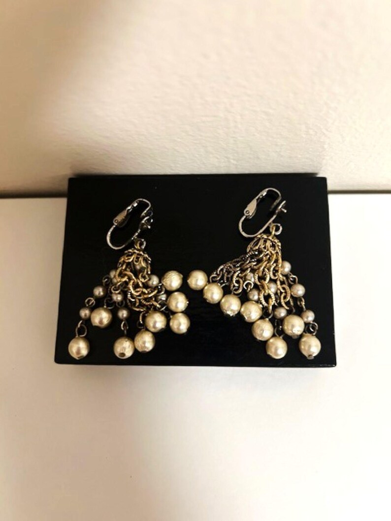 May include: A pair of dangling earrings with a mix of gold-toned metal and faux pearls. The earrings feature a cluster of small pearls hanging from a decorative metal design. The earrings are displayed on a black surface.