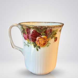 Puede incluir: Taza de cerámica blanca con un diseño floral de rosas en tonos rojos, amarillos y rosas. La taza tiene un borde y un asa dorados. La taza tiene estrías verticales.