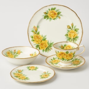 Könnte beinhalten: Ein cremefarbenes Porzellan-Teeservice mit gelben Rosenmustern. Das Set beinhaltet eine Tasse mit Untertasse, eine Schüssel, einen kleinen Teller und einen größeren Teller. Jedes Teil hat einen goldenen Rand.