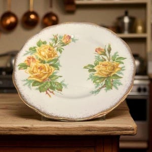 May include: A white porcelain plate with a scalloped edge and gold trim. The plate features a floral design of yellow roses and green leaves. The plate is displayed on a wooden surface.