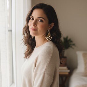 May include: A woman wearing a cream-colored sweater and gold earrings with pearl accents. The earrings are a prominent feature, adding a touch of elegance. The woman is near a window with sheer curtains, and a bed is visible in the background.