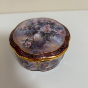 May include: A round trinket box with a floral design on the lid. The lid and sides have a burgundy border with gold trim. The box is painted with a bouquet of flowers in a vase.