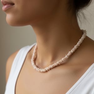 May include: A light pink shell necklace. The necklace is made of small, irregular shell pieces strung together, creating a textured, organic appearance. The necklace is worn against a white vest top.