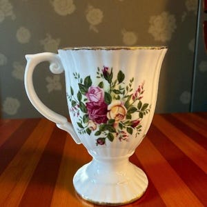 Peut inclure: Tasse à thé en porcelaine blanche avec une anse et un piedestal. La tasse est ornée d'un motif floral de roses roses, rouges et jaunes avec des feuilles vertes. Le bord et la base ont une bordure dorée. La tasse est sur une surface en bois rayé.