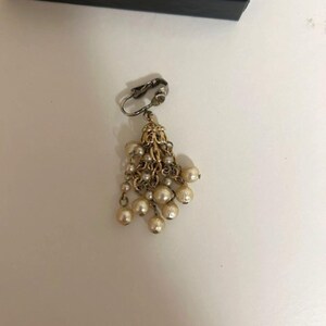 May include: A vintage clip-on earring with a cluster of faux pearls. The earring features a silver-tone clip and a gold-tone filigree design. Several strands of small pearls cascade from the filigree, creating a dangling effect. The pearls are off-white.