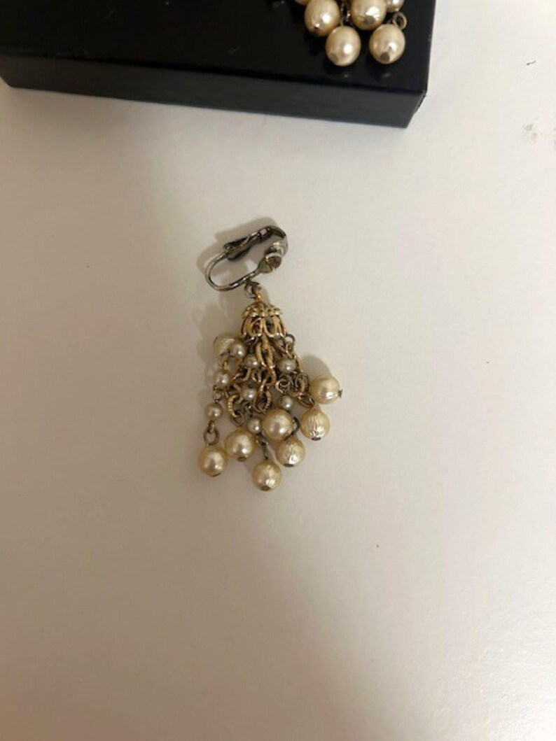 May include: A vintage clip-on earring with a cluster of faux pearls. The earring features a silver-tone clip and a gold-tone filigree design. Several strands of small pearls cascade from the filigree, creating a dangling effect. The pearls are off-white.