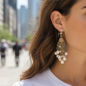May include: Gold and silver-toned chandelier earrings with dangling chains and faux pearls. The earrings have a decorative top with a small crystal accent. The earrings are worn by a person with long, wavy brown hair.