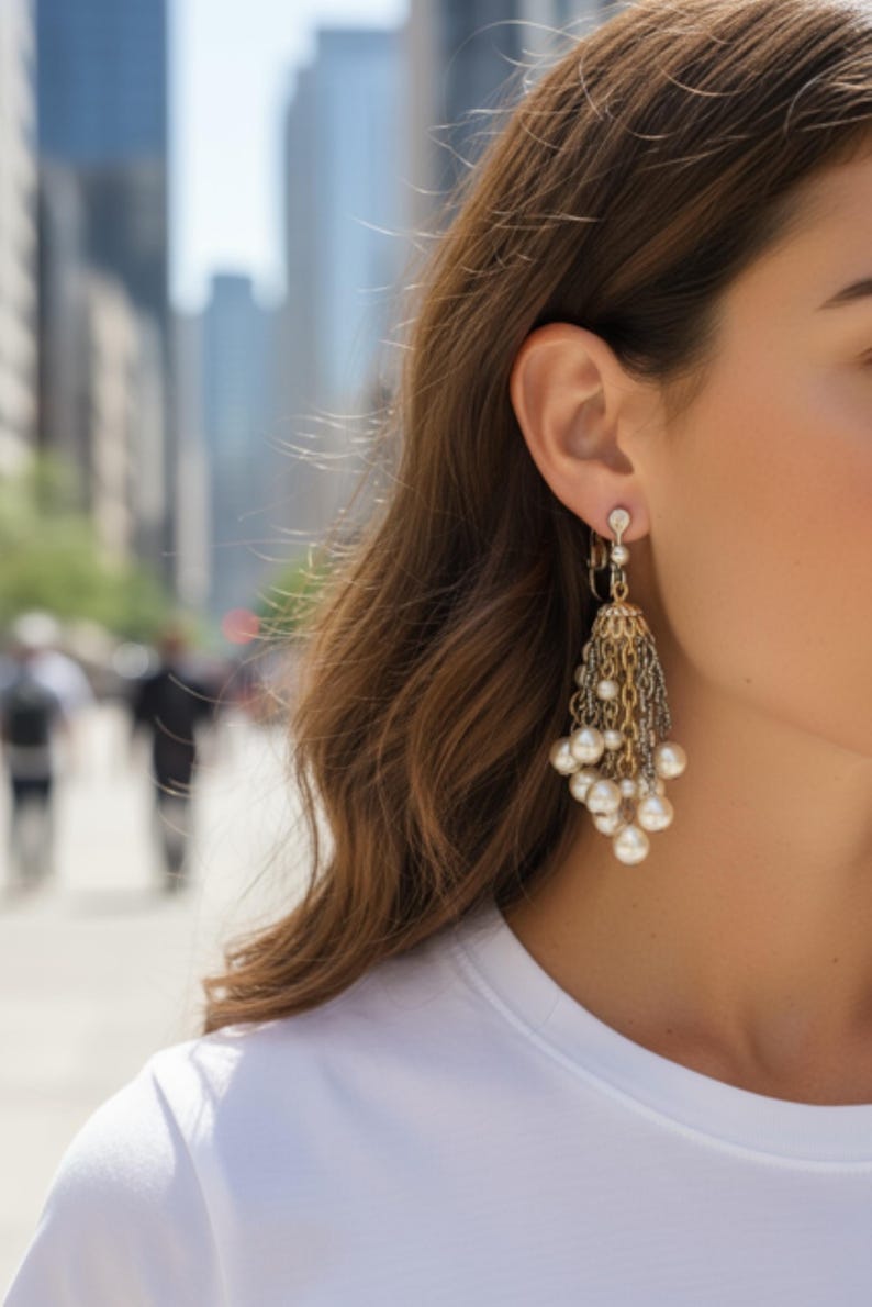 May include: Gold and silver-toned chandelier earrings with dangling chains and faux pearls. The earrings have a decorative top with a small crystal accent. The earrings are worn by a person with long, wavy brown hair.