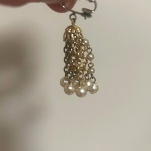 May include: A gold-toned earring with a dangling tassel design. The top portion features a filigree cap, with multiple strands of gold chains and small, round, off-white faux pearls at the ends. The earring is held by a silver-colored clip.