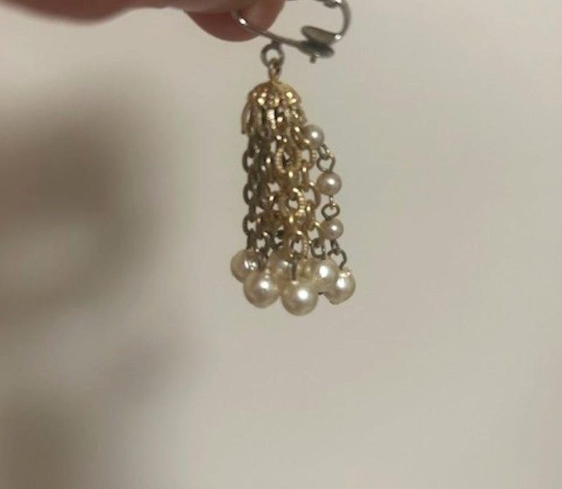 May include: A gold-toned earring with a dangling tassel design. The top portion features a filigree cap, with multiple strands of gold chains and small, round, off-white faux pearls at the ends. The earring is held by a silver-colored clip.