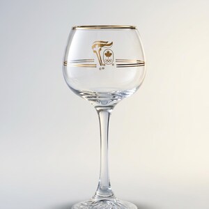 May include: A clear wine glass with a gold rim and gold Olympic logo. The logo features a torch, maple leaf, and Olympic rings. The glass has a long stem and a decorative base.
