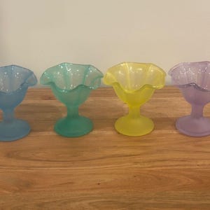 May include: Four frosted glass dessert dishes in pastel colors: blue, green, yellow, and purple. Each dish has a ruffled rim and a pedestal base, perfect for serving ice cream or other treats.