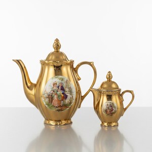May include: A gold-coloured porcelain teapot and a matching smaller creamer, both featuring a glossy metallic finish, a scalloped lid with a finial, and a central oval medallion depicting a colourful illustration of two figures in a garden setting.