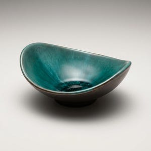 May include: A teal ceramic bowl with a dark brown rim and base. The bowl has an unusual, asymmetrical shape, with a smooth, glossy finish. The interior of the bowl has a darker, mottled appearance.