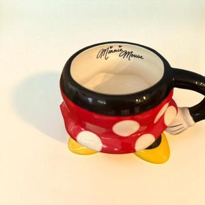 May include: A ceramic Minnie Mouse mug with a black handle and rim. The mug is red with white polka dots and yellow shoes. The words "Minnie Mouse" are written in black script inside the mug.